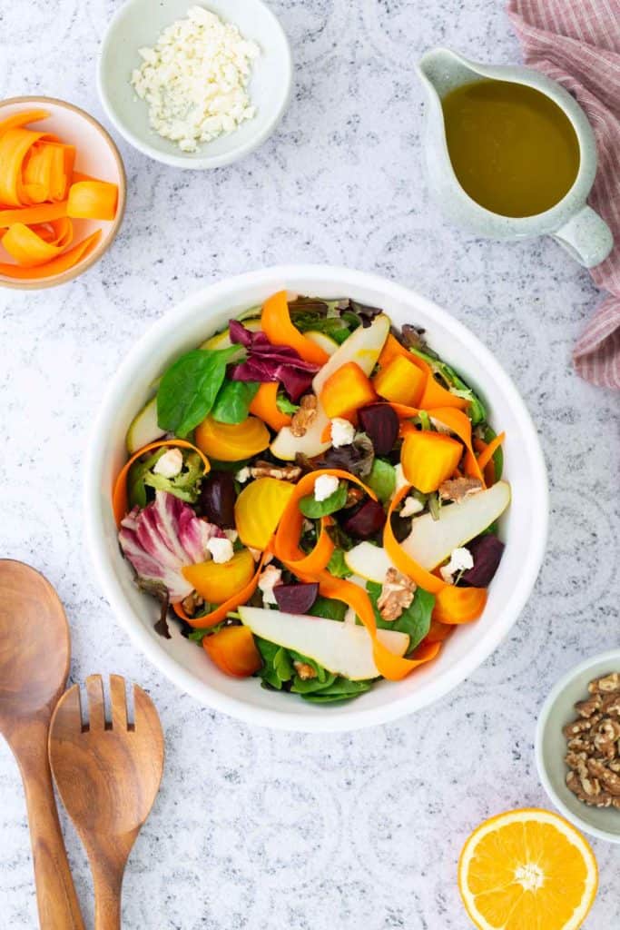 A bowl of mixed salad with greens, sliced carrots, beets, walnuts, cheese, and pear, surrounded by dressing, cheese, carrot ribbons, walnuts, and a wooden serving set.
