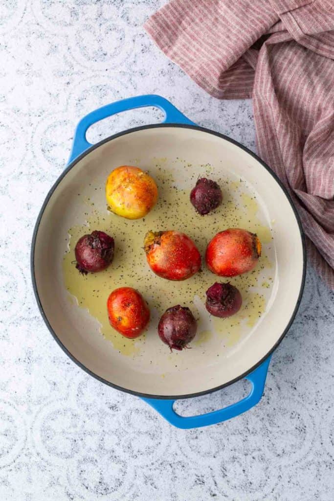 A blue-handled baking dish with whole red and yellow beets, drizzled with oil and sprinkled with pepper, on a light textured surface next to a pink-striped cloth.