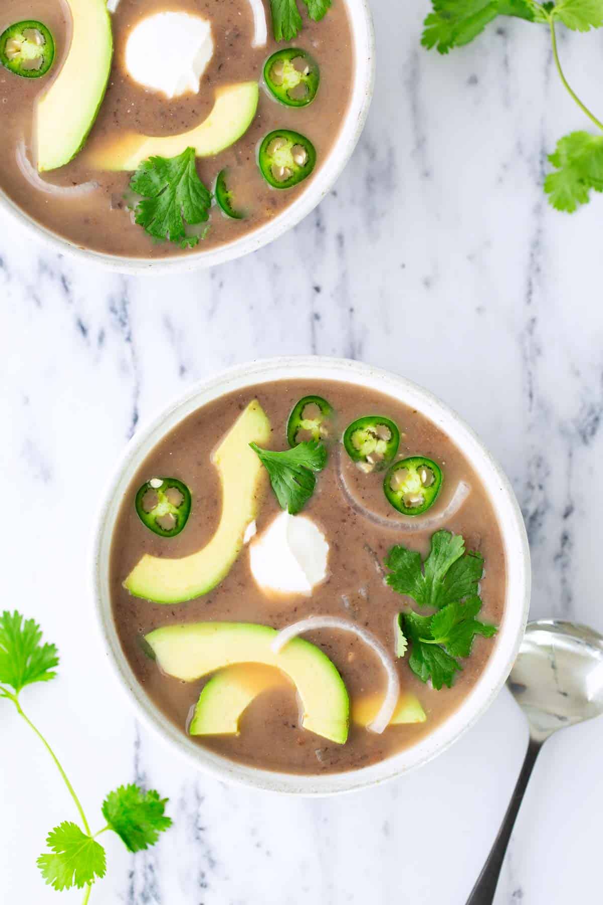 Two bowls of black bean soup topped with avocado slices, jalapeño rounds, sour cream, and cilantro on a white marble surface with scattered cilantro leaves and a spoon.