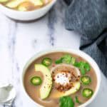 Two bowls of creamy soup topped with avocado slices, jalapeño, cilantro, sour cream, and red pepper flakes, with a spoon and a striped napkin nearby on a marble surface.