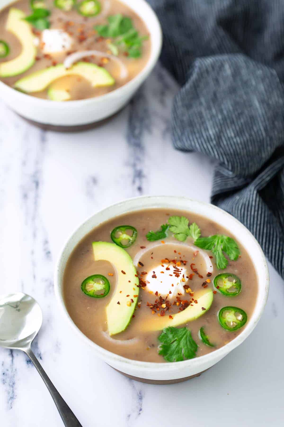 Two bowls of creamy soup topped with avocado slices, jalapeño, cilantro, sour cream, and red pepper flakes, with a spoon and a striped napkin nearby on a marble surface.