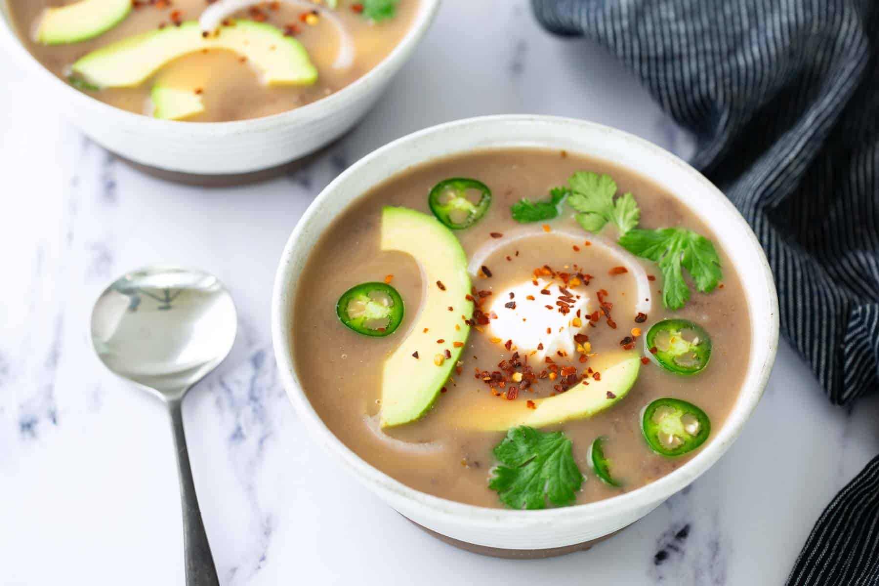 A bowl of creamy soup topped with avocado slices, jalapeño slices, cilantro, a dollop of cream, and red pepper flakes. A spoon and striped napkin are beside the bowl.