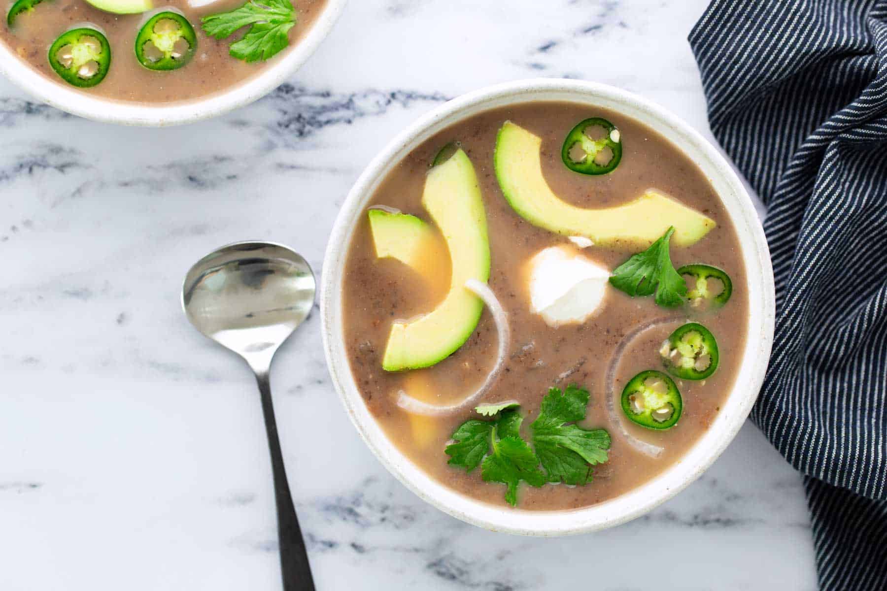 A bowl of black bean soup garnished with avocado slices, jalapeño, onion rings, cilantro, and sour cream sits on a marble surface next to a spoon and a striped cloth.