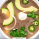 A bowl of creamy black bean soup topped with avocado slices, jalapeño, cilantro, onion, and a dollop of sour cream.