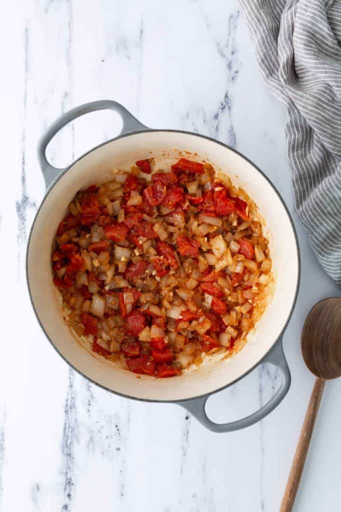 A large pot on a marble surface containing sautéed onions and diced tomatoes, with a wooden spoon and striped towel next to it.