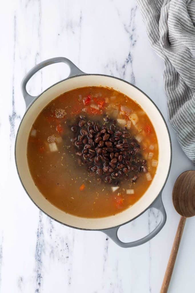 A pot of black bean soup with diced vegetables and broth sits on a marble surface next to a striped towel and a wooden spoon.
