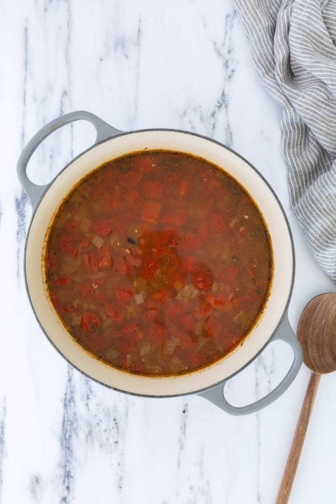 A pot of tomato-based soup with visible chopped tomatoes and onions sits on a marble surface beside a wooden spoon and a striped kitchen towel.