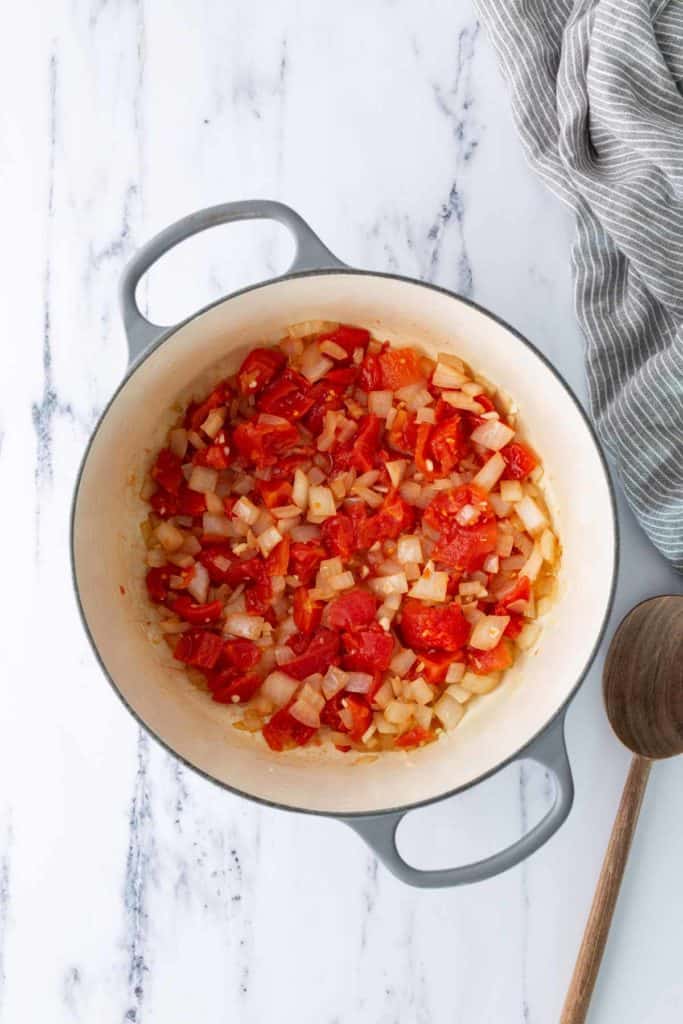 Diced tomatoes and onions cooking in a white pot on a marble surface, with a wooden spoon and a striped cloth nearby.