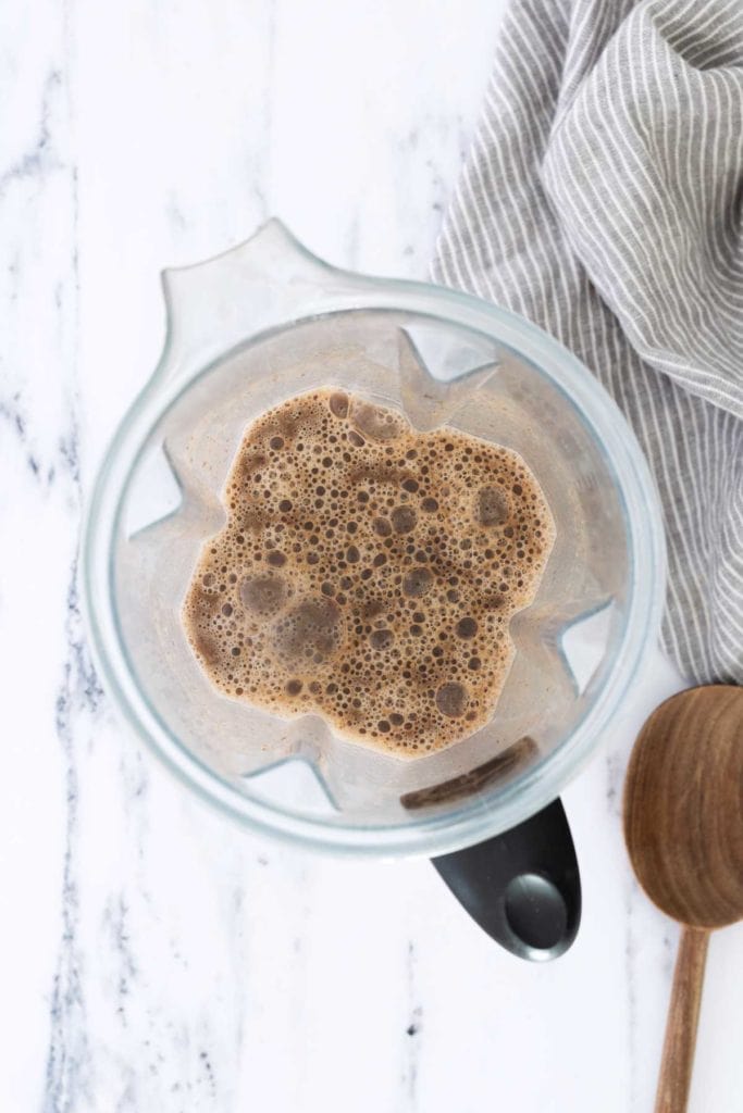 A top-down view of a blender filled with a frothy brown liquid, next to a wooden spoon and a gray striped cloth on a marble surface.