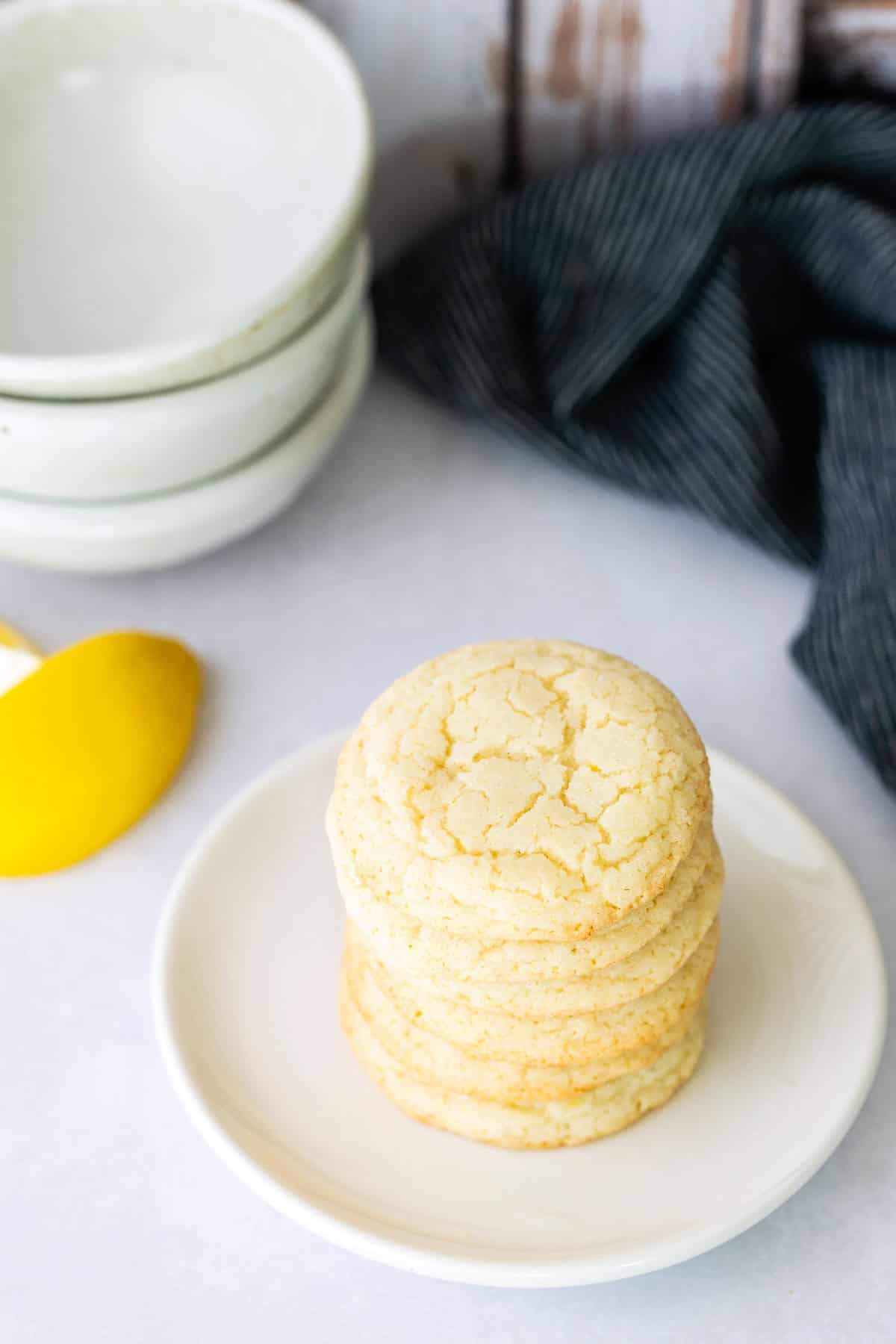 A stack of sugar cookies on a white plate, with a stack of white bowls, a striped cloth, and a lemon in the background.