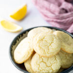 A bowl of round, cracked lemon cookies sits on a white surface with lemon wedges and a purple cloth in the background.