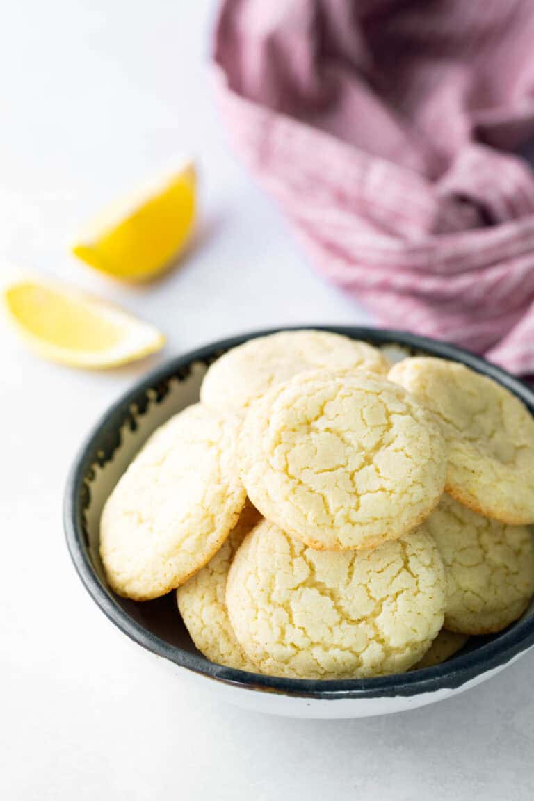 A bowl of round, cracked lemon cookies sits on a white surface with lemon wedges and a purple cloth in the background.