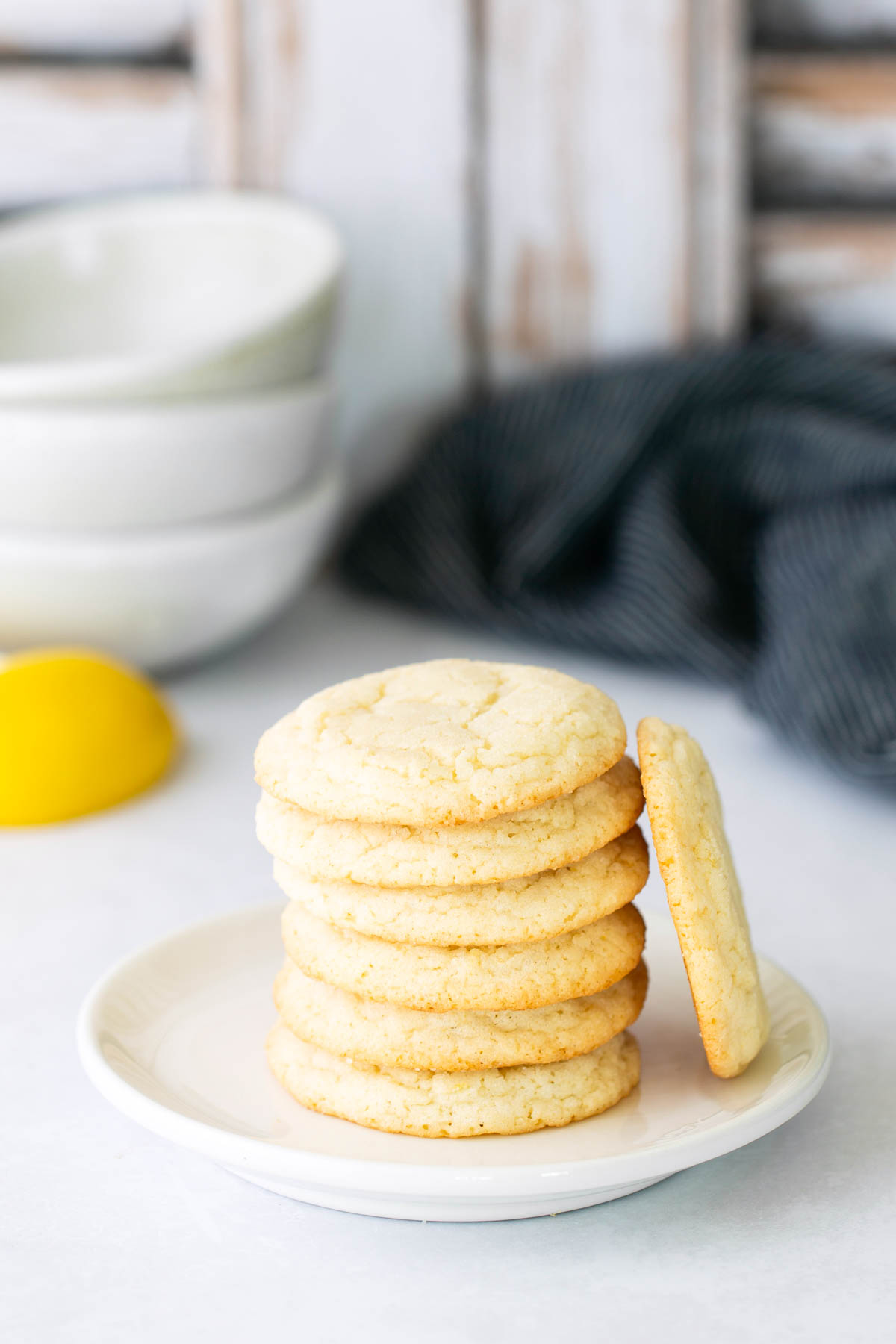A stack of sugar cookies sits on a white plate, with one cookie leaning against the stack; bowls and a lemon are in the background.