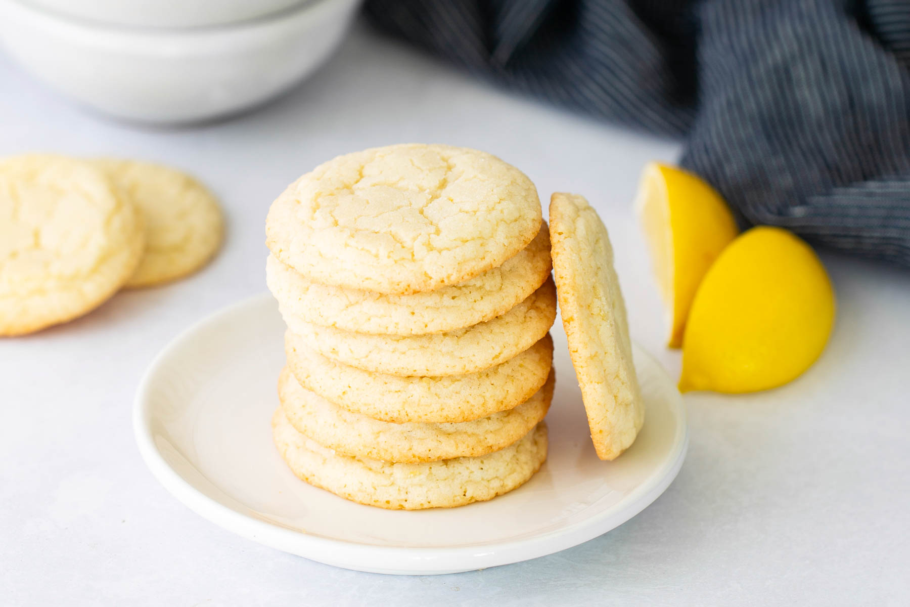A stack of sugar cookies on a white plate, with two lemon wedges and additional cookies in the background.
