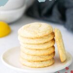 A stack of six lemon cookies is displayed on a white plate, with one cookie propped up on the side. The background shows a lemon and blurred kitchen items. Text reads "LEMON COOKIES.