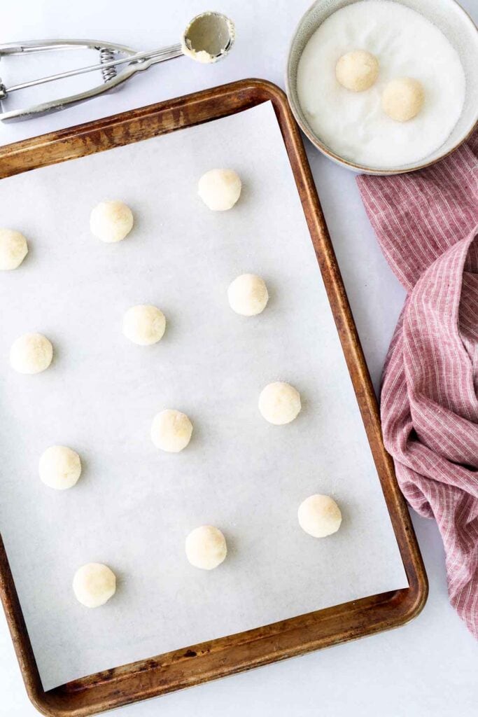 A baking sheet lined with parchment paper holds evenly spaced raw cookie dough balls. Nearby are a bowl of sugar with two dough balls, a scoop, and a pink cloth.