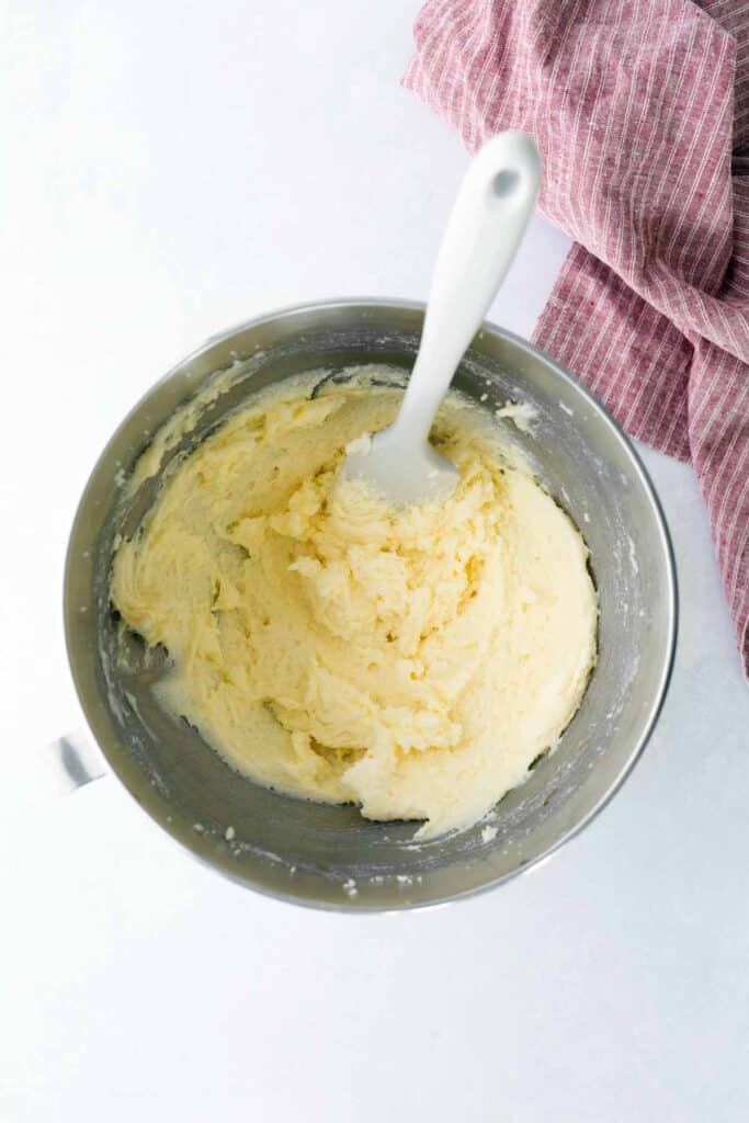A mixing bowl filled with creamy, pale yellow cake batter and a white spatula, with a pink striped kitchen towel beside it.