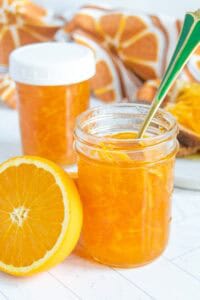 A glass jar of orange marmalade with a spoon, a halved orange, and another jar in the background on a white surface.