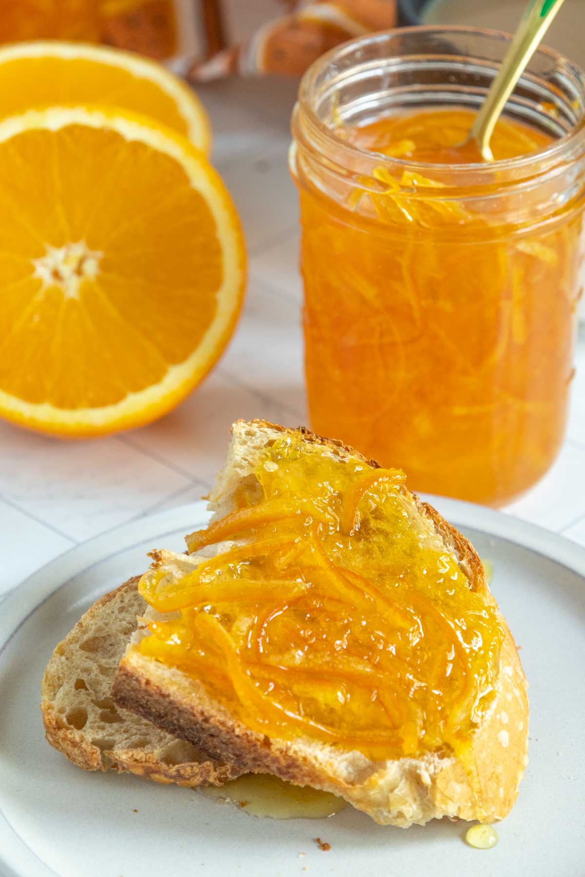 A slice of bread with orange marmalade on a plate, a jar of marmalade with a spoon, and two orange slices in the background.