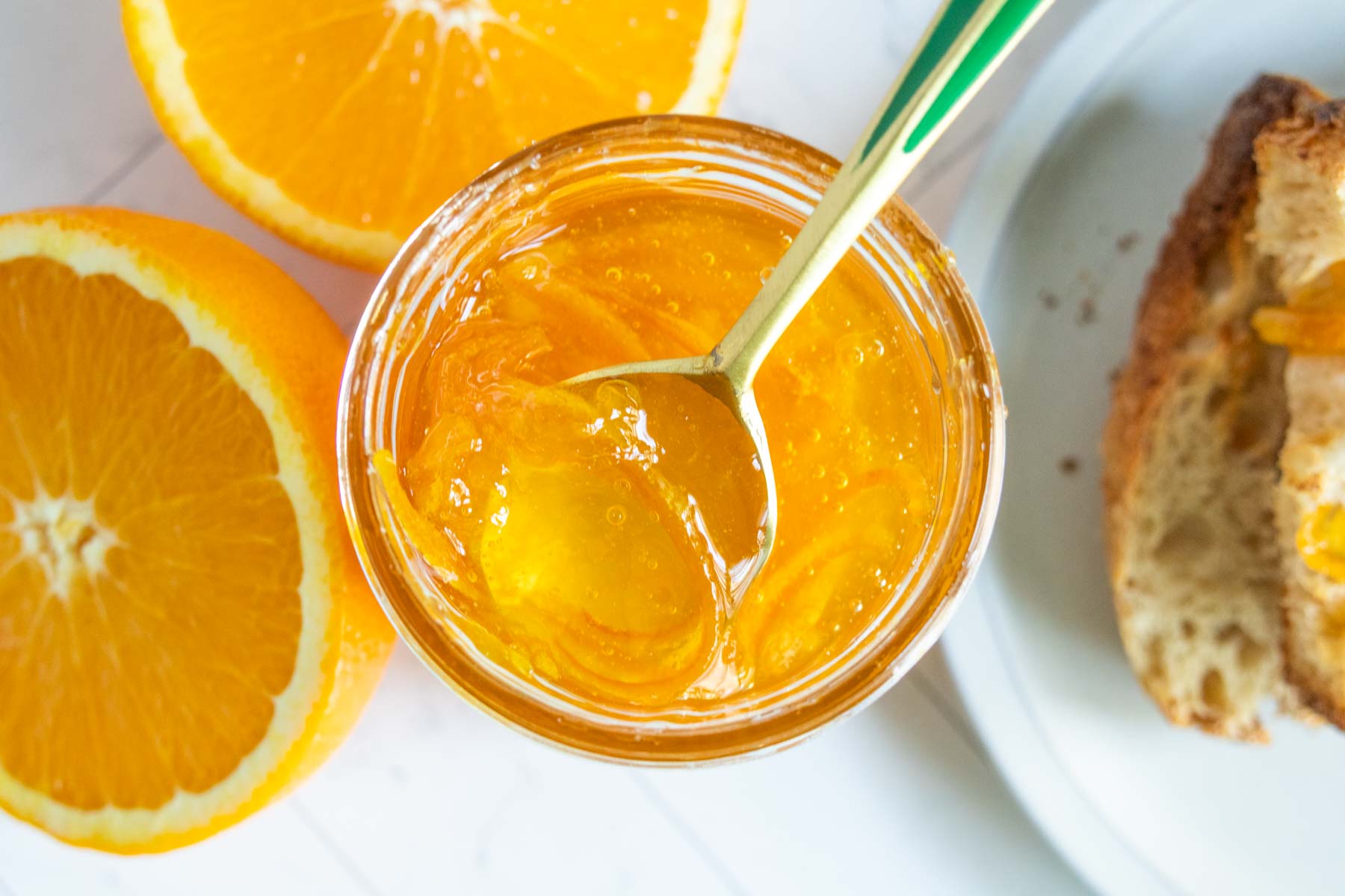 A jar of orange marmalade with a spoon inside, next to halved oranges and a slice of bread topped with marmalade.