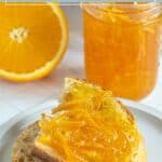 A slice of bread topped with orange marmalade sits on a plate, with a jar of marmalade and a halved orange in the background.