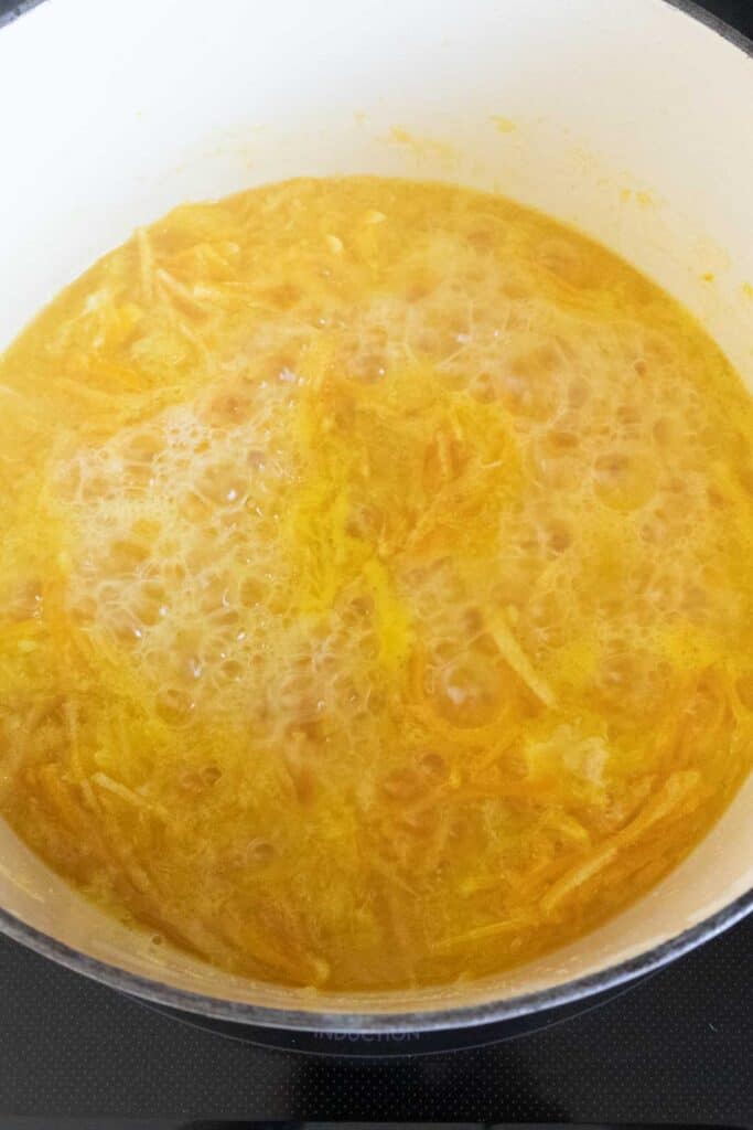 A pot of orange marmalade is boiling, with visible bubbles and shredded orange peel in the mixture.