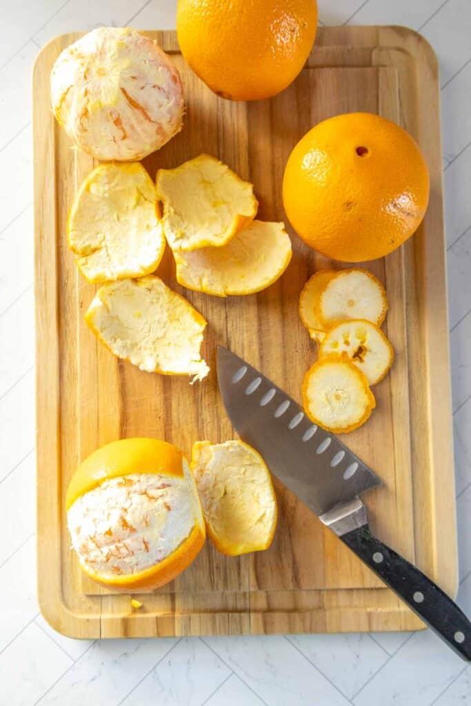 A cutting board with whole, partially peeled, and sliced oranges, orange peels, and a knife.