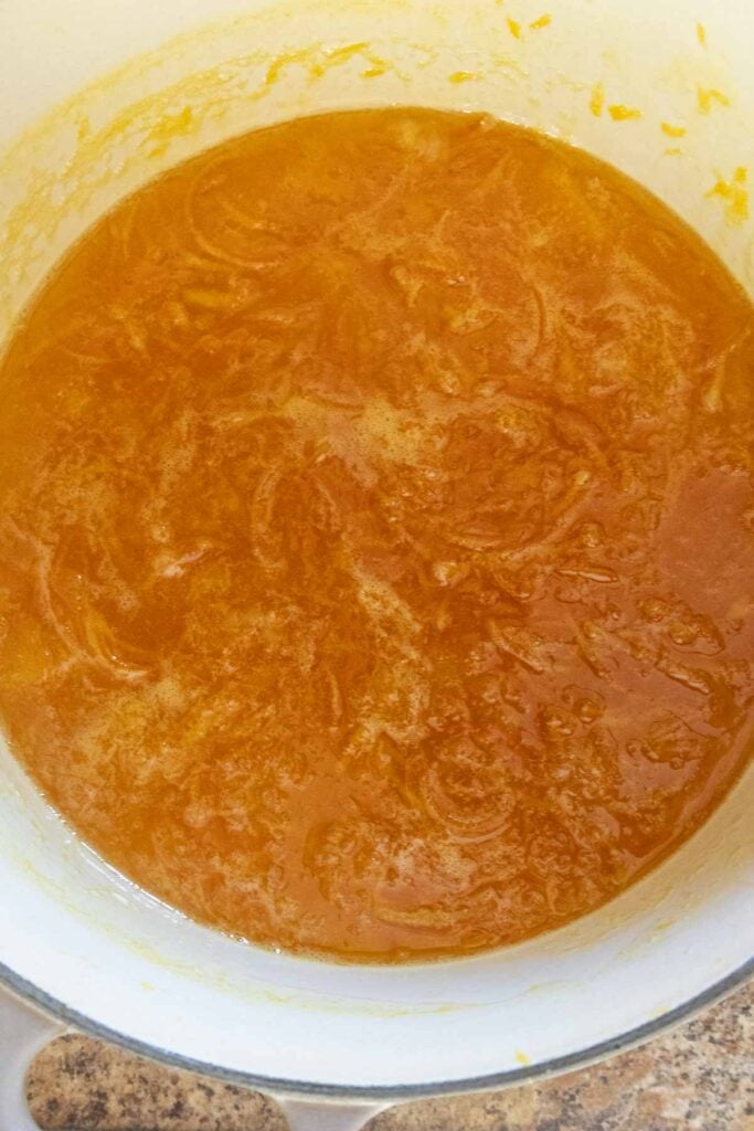A pot filled with orange marmalade mixture in the process of cooking, showing visible orange peel strips and bubbling liquid.