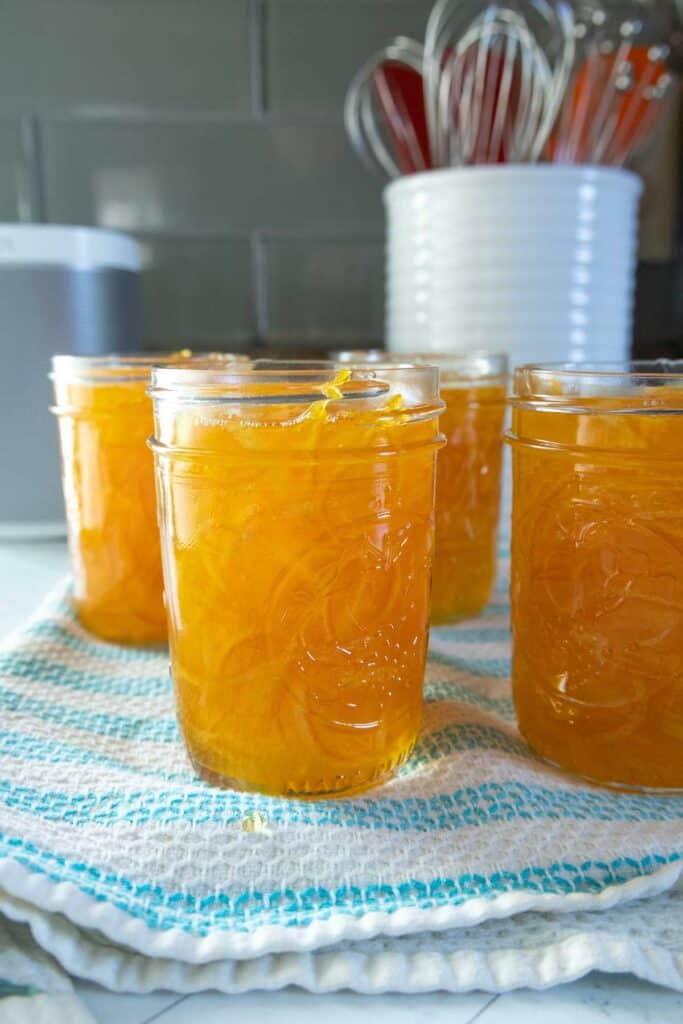 Four glass jars filled with orange marmalade sit on a blue and white dish towel, with kitchen utensils and stacked bowls in the blurred background.