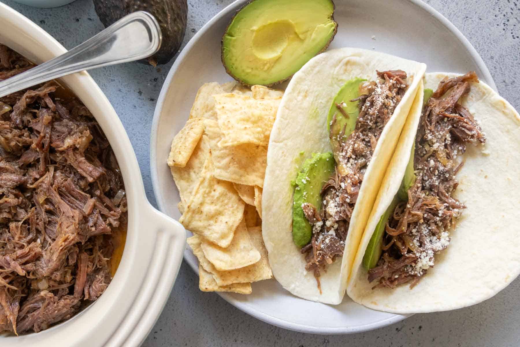 Two flour tortillas filled with shredded beef, avocado slices, and cheese on a plate with tortilla chips and half an avocado, next to a dish of shredded beef.