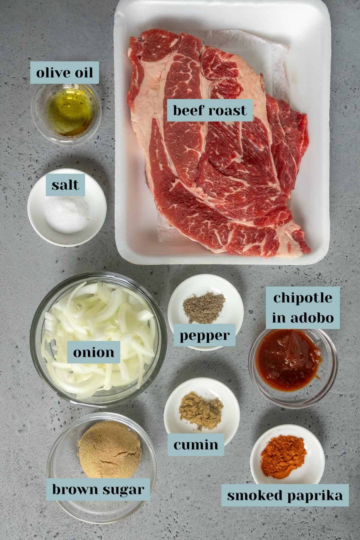 Raw beef roast on a tray surrounded by bowls containing olive oil, salt, sliced onion, brown sugar, pepper, cumin, chipotle in adobo, and smoked paprika on a gray surface.