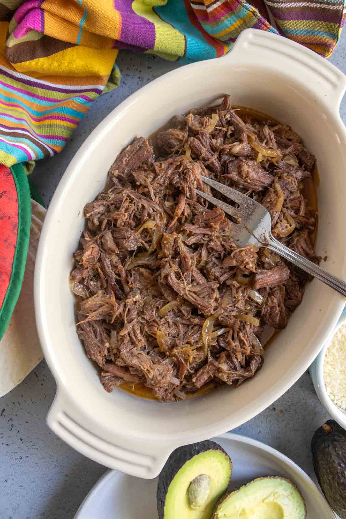 Shredded beef with onions in an oval white dish, served with a fork, surrounded by colorful cloth, avocado, tortillas, and rice.