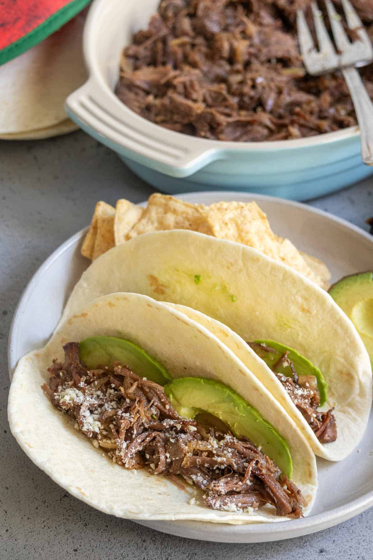 Two soft tacos filled with shredded beef, sliced avocado, and grated cheese are served on a plate with tortilla chips and more shredded beef in a dish in the background.