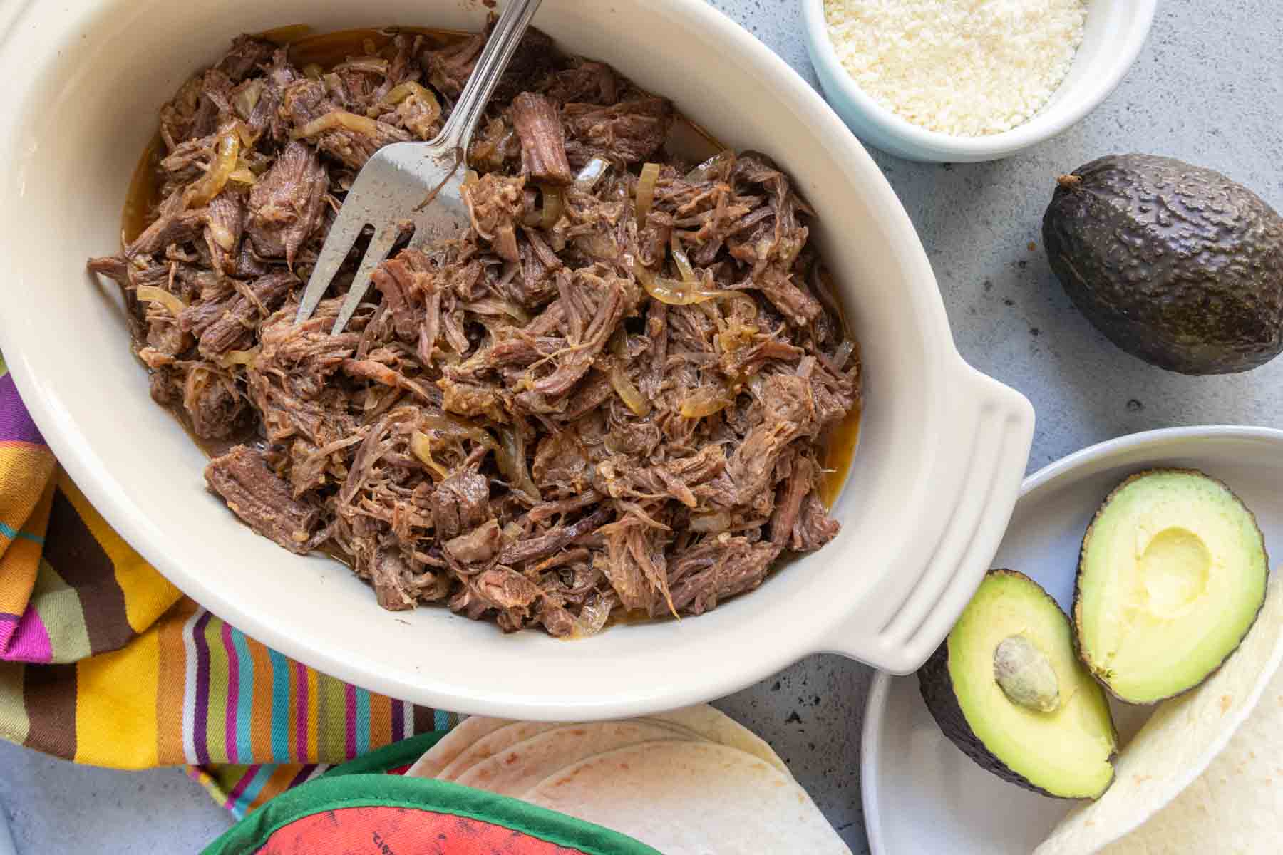 Shredded beef with onions in a white dish, surrounded by tortillas, sliced avocado, whole avocado, grated cheese, and a striped towel.