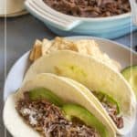 Two shredded beef tacos with sliced avocado and cheese on flour tortillas, served on a plate with tortilla chips. A dish of shredded beef is visible in the background.