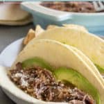 Two shredded beef tacos with avocado slices in flour tortillas, topped with grated cheese, served on a white plate.