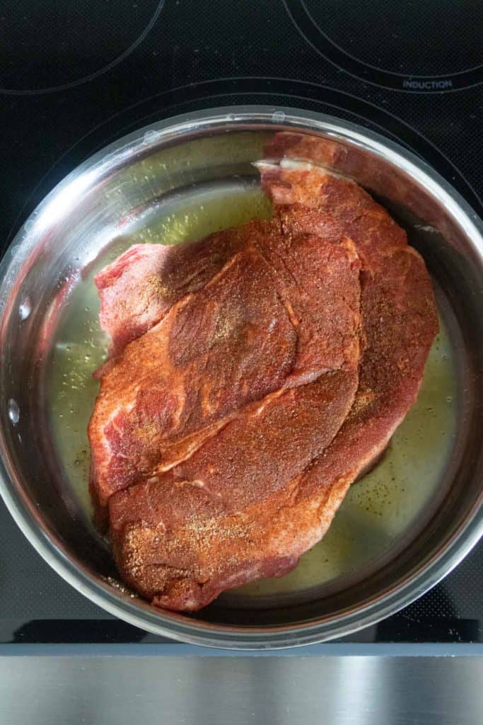 A large piece of seasoned raw meat sits in a stainless steel pan on a stovetop, ready to be cooked.
