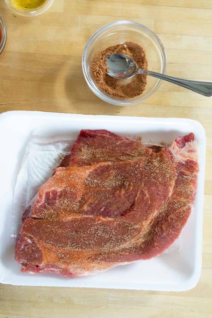 Raw pork ribs in a white tray topped with dry seasoning rub, with a bowl of extra dry rub and a spoon beside it on a wooden surface.