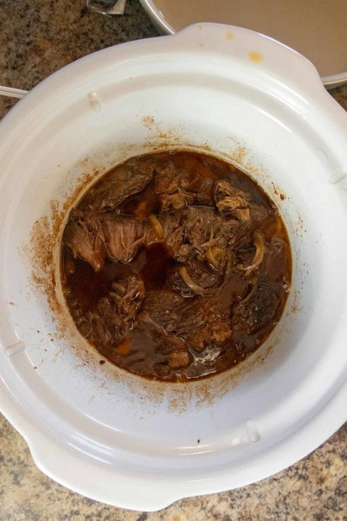 Chunks of cooked beef in a rich brown sauce inside a white slow cooker on a kitchen countertop.