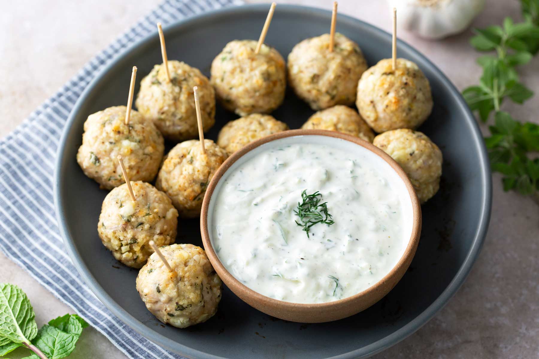 A plate of meatballs with toothpicks arranged around a bowl of creamy white dipping sauce topped with herbs.