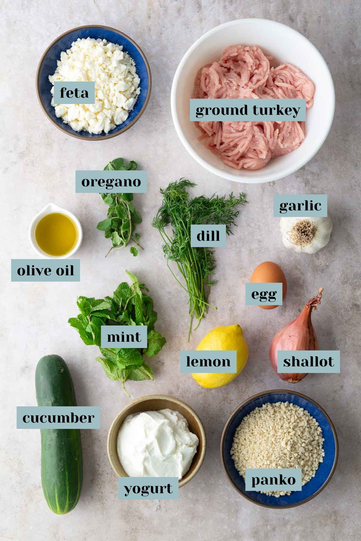 A flat lay of ingredients for a recipe including ground turkey, feta, olive oil, oregano, dill, garlic, egg, shallot, lemon, mint, cucumber, yogurt, and panko in separate bowls.