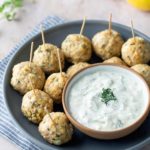 A plate of baked meatballs with toothpicks, served alongside a bowl of creamy white dipping sauce, garnished with herbs.