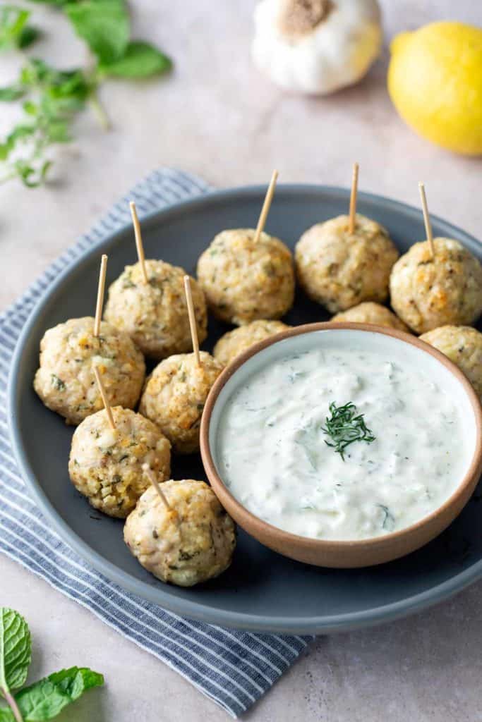 Greek Turkey Meatballs A plate of baked meatballs with toothpicks, served alongside a bowl of creamy white dipping sauce, garnished with herbs.