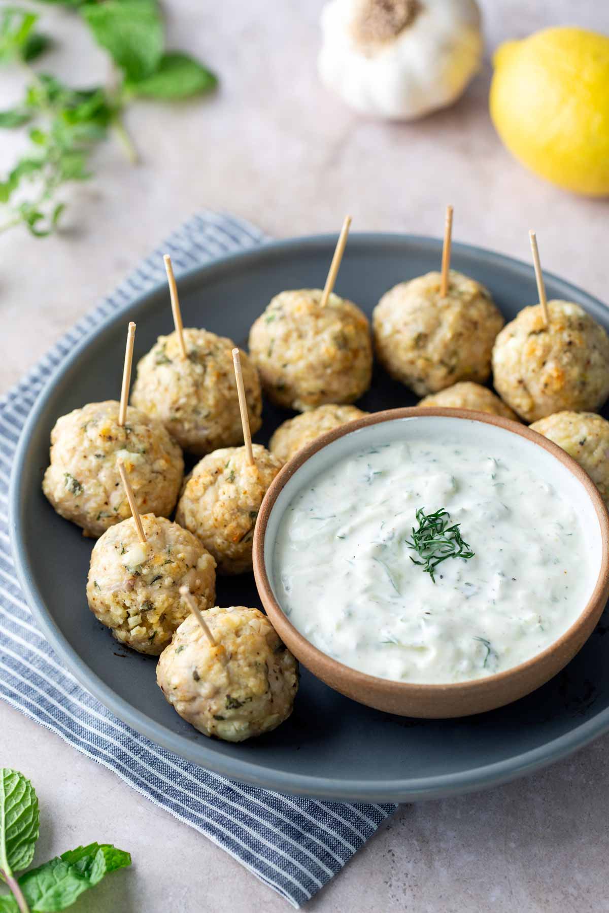 A plate of baked meatballs with toothpicks, served alongside a bowl of creamy white dipping sauce, garnished with herbs.