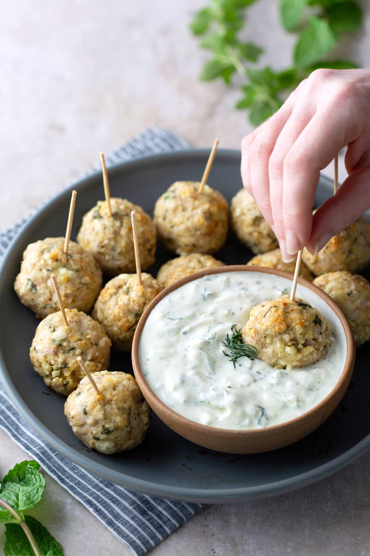 A hand dips a toothpick skewered meatball into a bowl of creamy white sauce, surrounded by more meatballs on a gray plate.