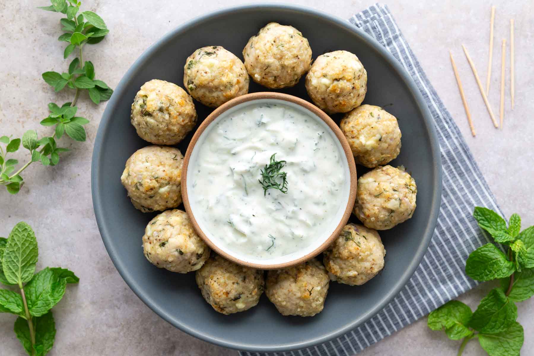 A plate of meatballs arranged in a circle around a bowl of white dipping sauce, garnished with herbs, on a gray plate with fresh mint and striped napkin nearby.