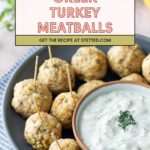 A plate of Greek turkey meatballs with toothpicks, served with a bowl of creamy dipping sauce, displayed on a blue plate.