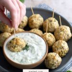 A hand dips a turkey meatball on a toothpick into a bowl of white sauce, surrounded by more meatballs on a gray plate. Text overlay reads "greek turkey meatballs.