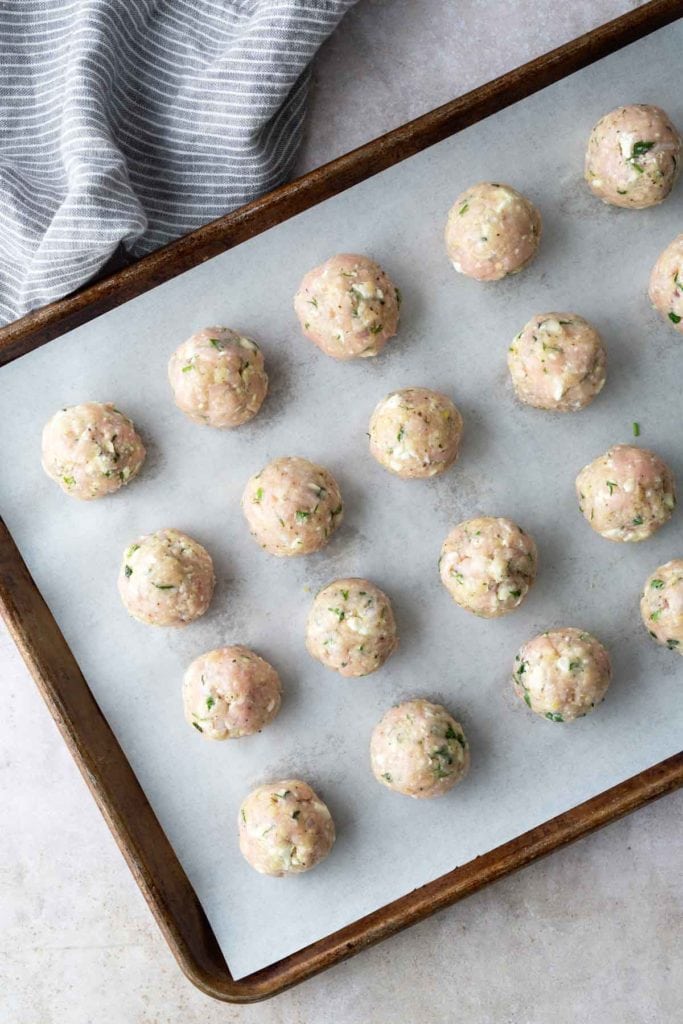 Uncooked meatballs arranged in rows on a parchment-lined baking sheet next to a striped cloth.