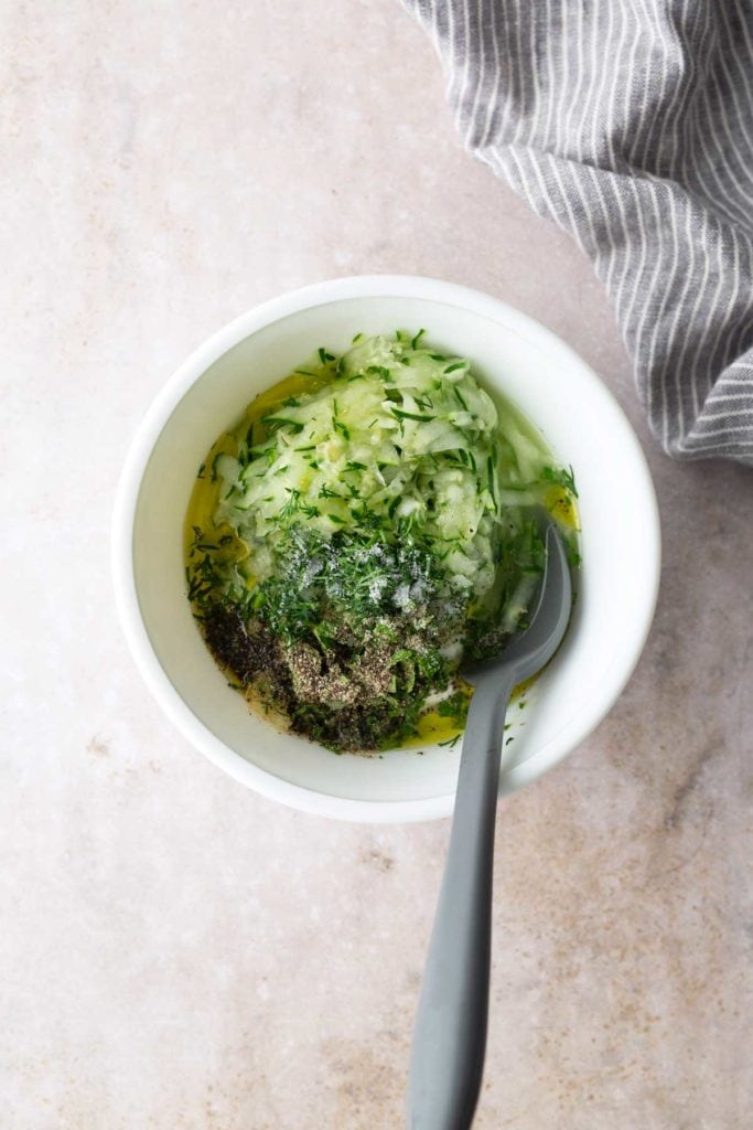 A white bowl containing grated cucumber, chopped dill, black pepper, salt, and olive oil with a gray spoon, placed on a light surface next to a striped cloth.